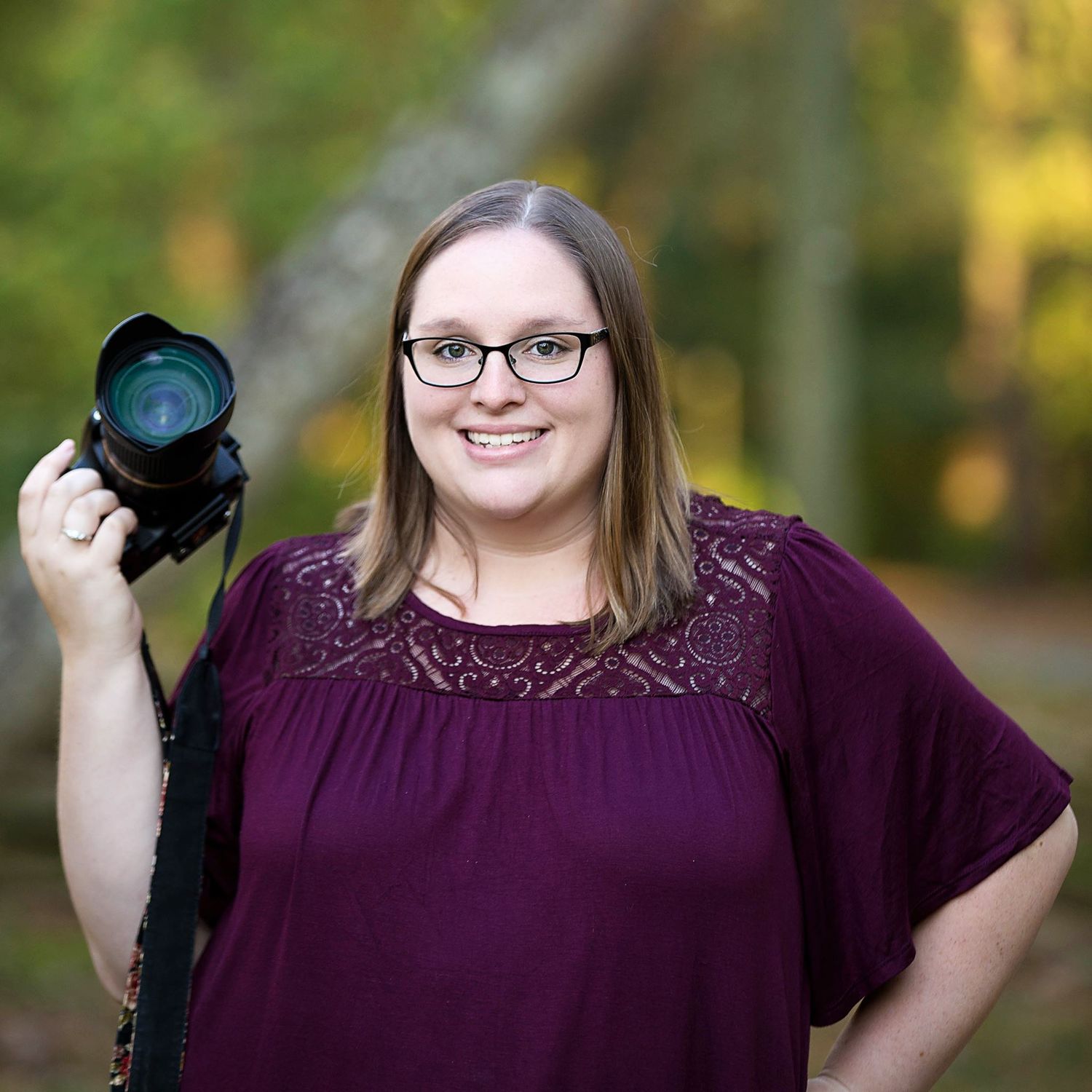 Professional photographer in burgundy lace top poses with camera equipment outdoors.