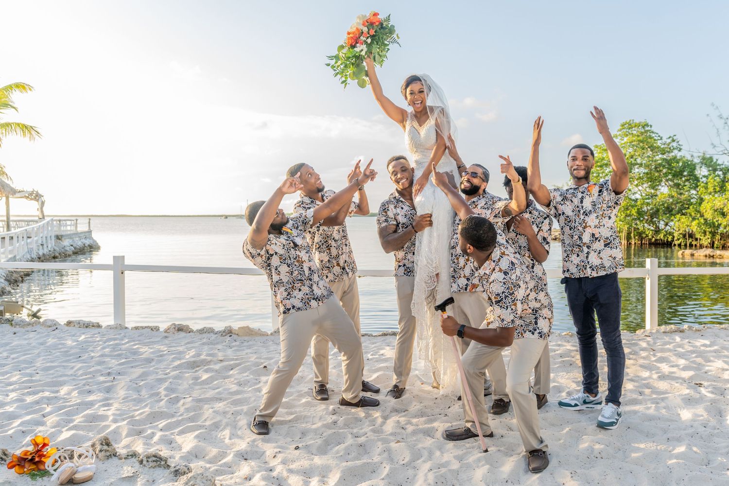 A joyous wedding party celebrates on a sunny beach with matching floral print shirts and a bouquet raised high in the air.