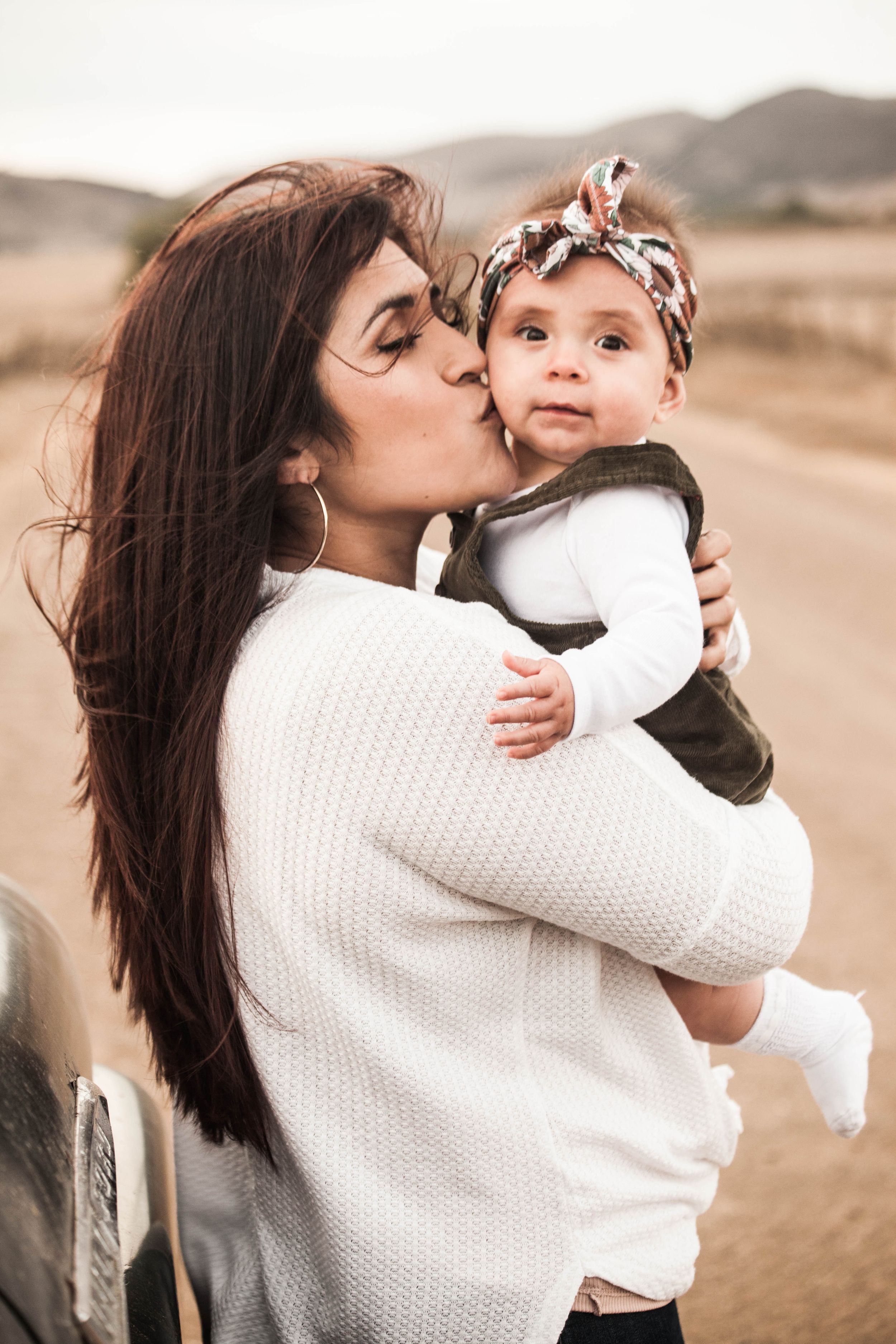 A tender moment between parent and baby outdoors in white sweater against a desert landscape background.