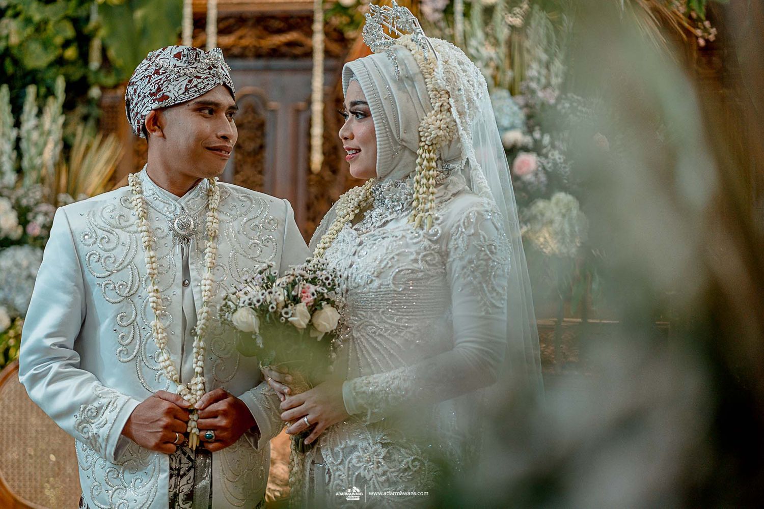 A traditional Indonesian wedding couple in elegant white and silver attire share an intimate moment.