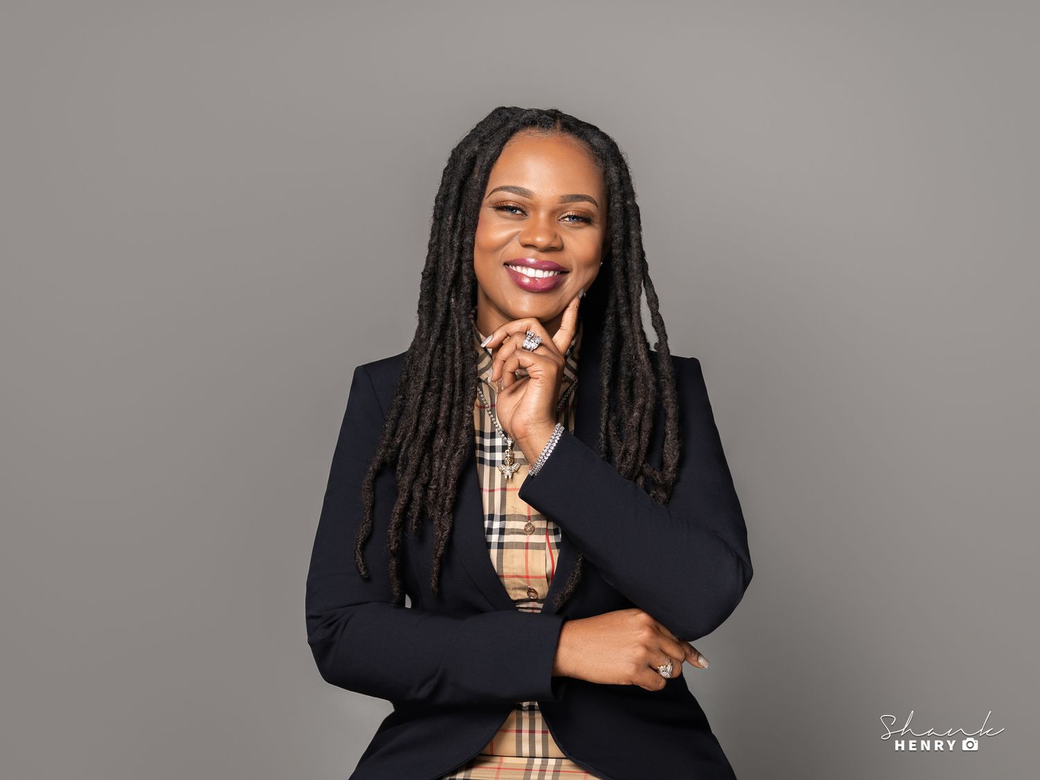 Professional headshot of woman in navy blazer and plaid shirt with long locs hairstyle against grey background.