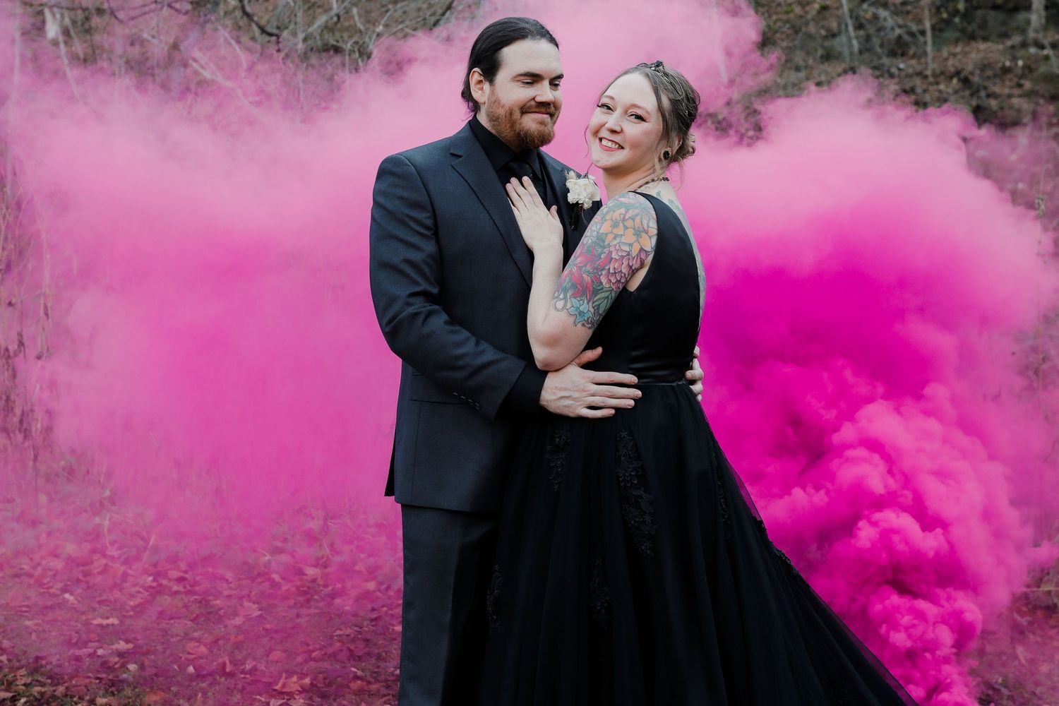 Couple in matching black outfits posing with vibrant pink smoke bombs in an outdoor wedding photo.