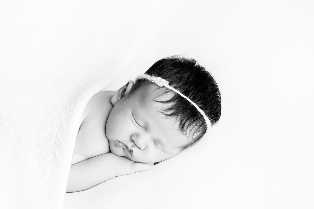 Black and white image of newborn baby girl laying on white blanket with hand resting under her cheek.