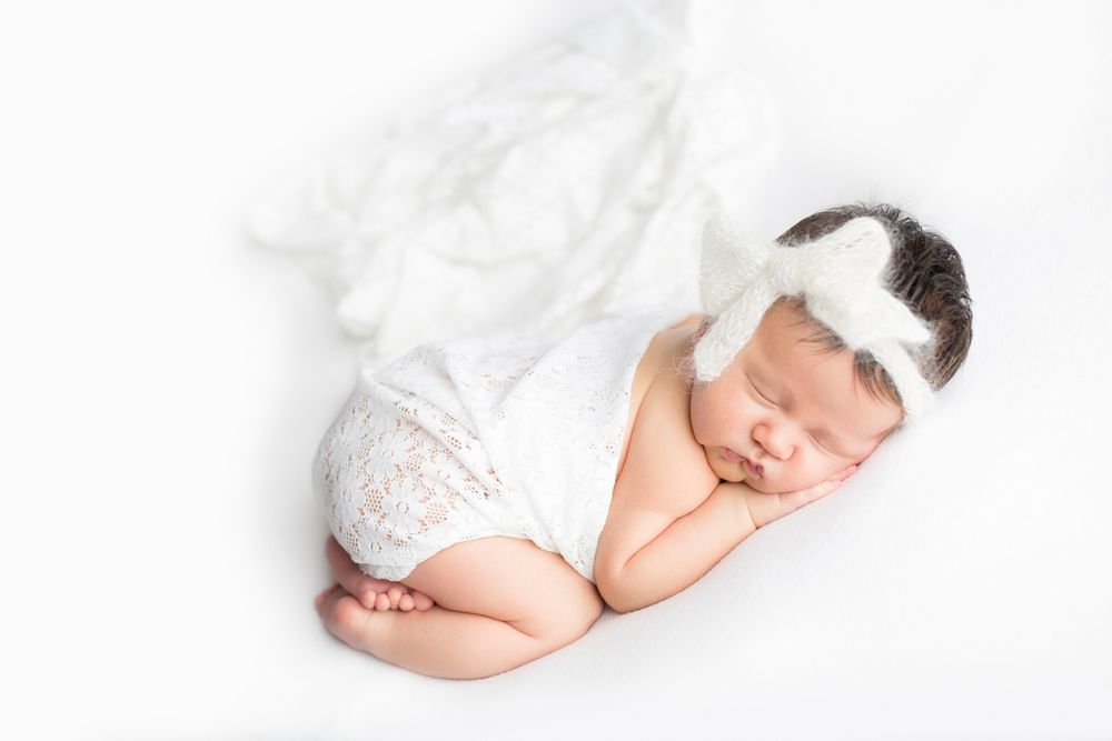 Newborn baby girl on a white blanket with a lace romper and white headband.