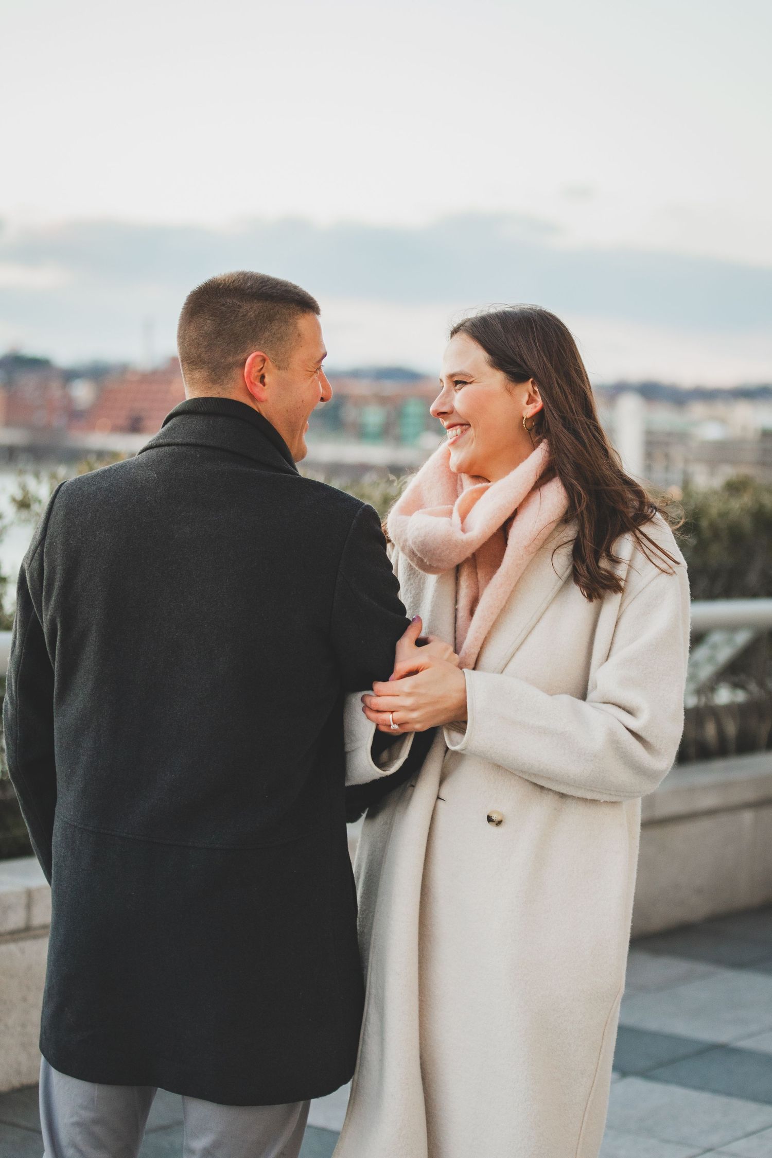 A couple shares a tender moment outdoors wearing winter coats against a city backdrop.