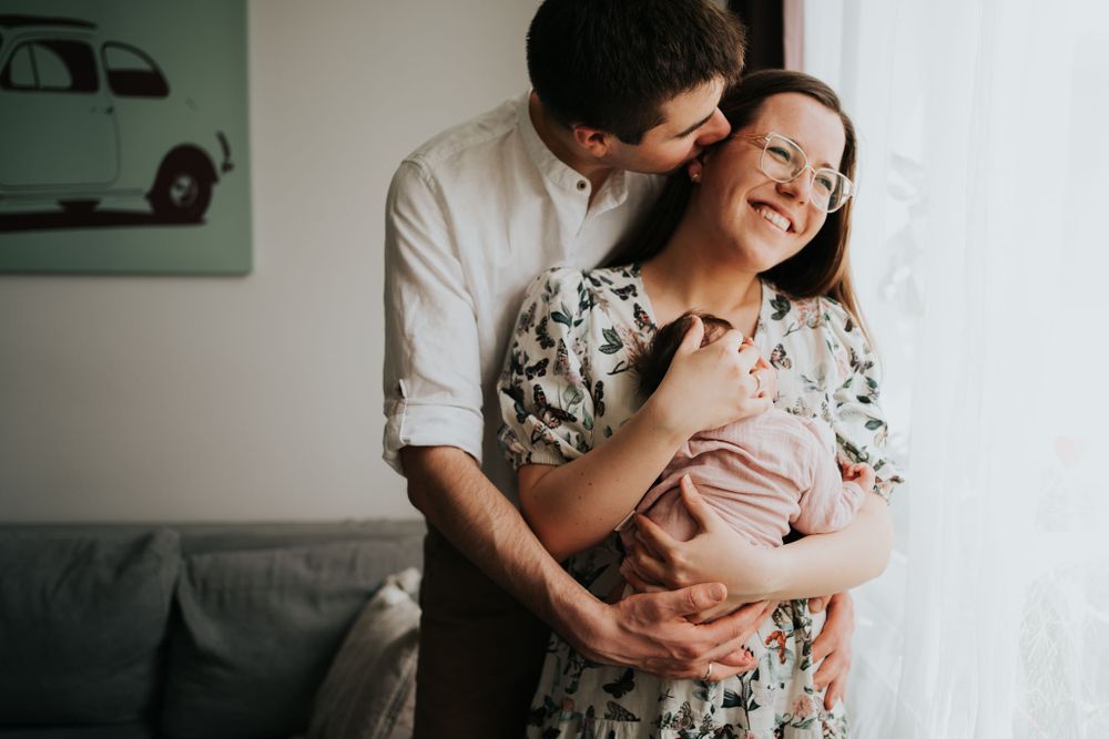 mama i tata stoją przy oknie i tulą noworodka - fotograf noworodkowy Kraków