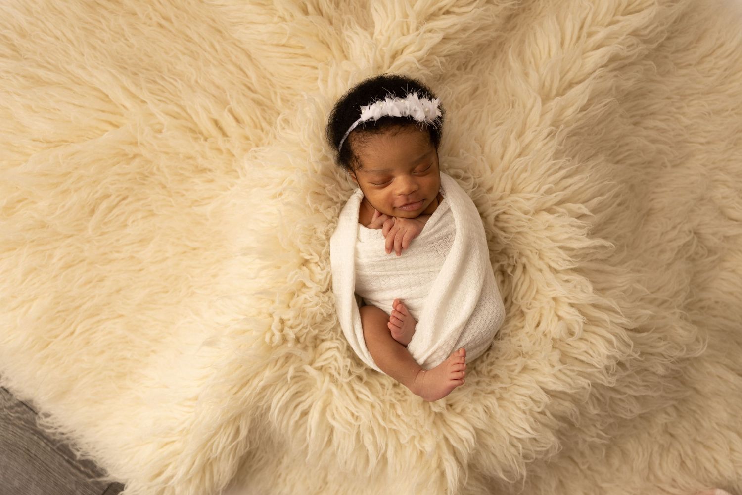 Newborn baby wrapped in white blanket sleeping on cream colored fluffy fur backdrop.
