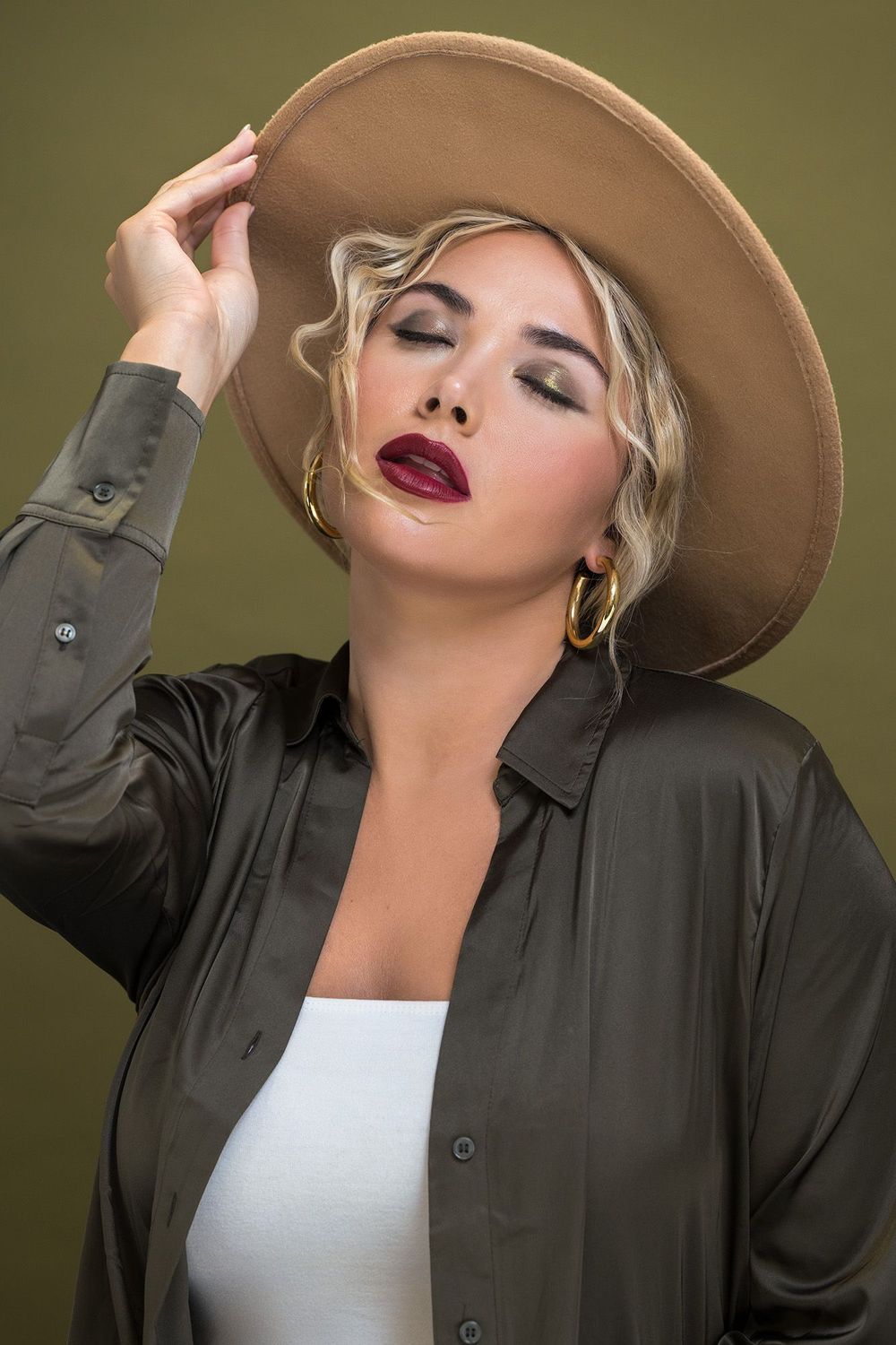 Blonde woman with bold red lips wearing a wide-brim hat and olive shirt with eyes closed.