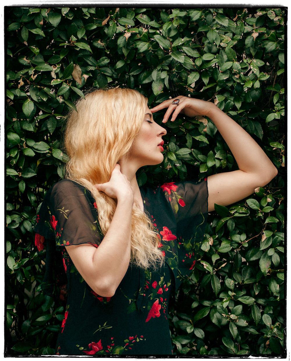 Blonde woman in floral dress posing against lush green ivy wall in natural light.