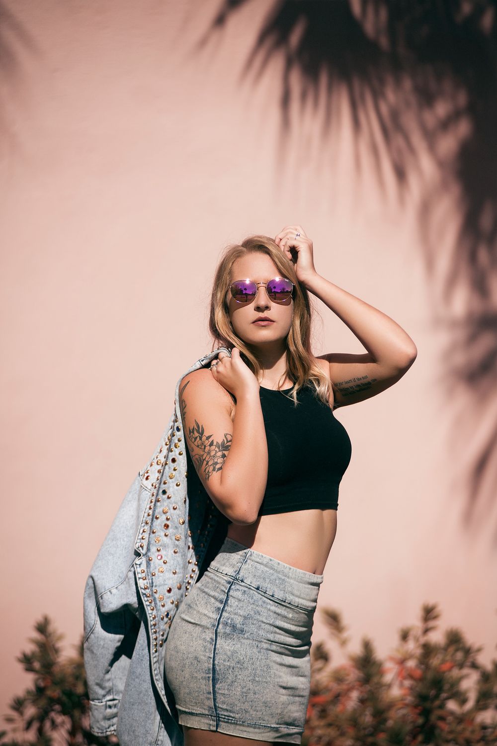 Blonde woman in crop top, denim skirt, and purple sunglasses posing against pink wall.
