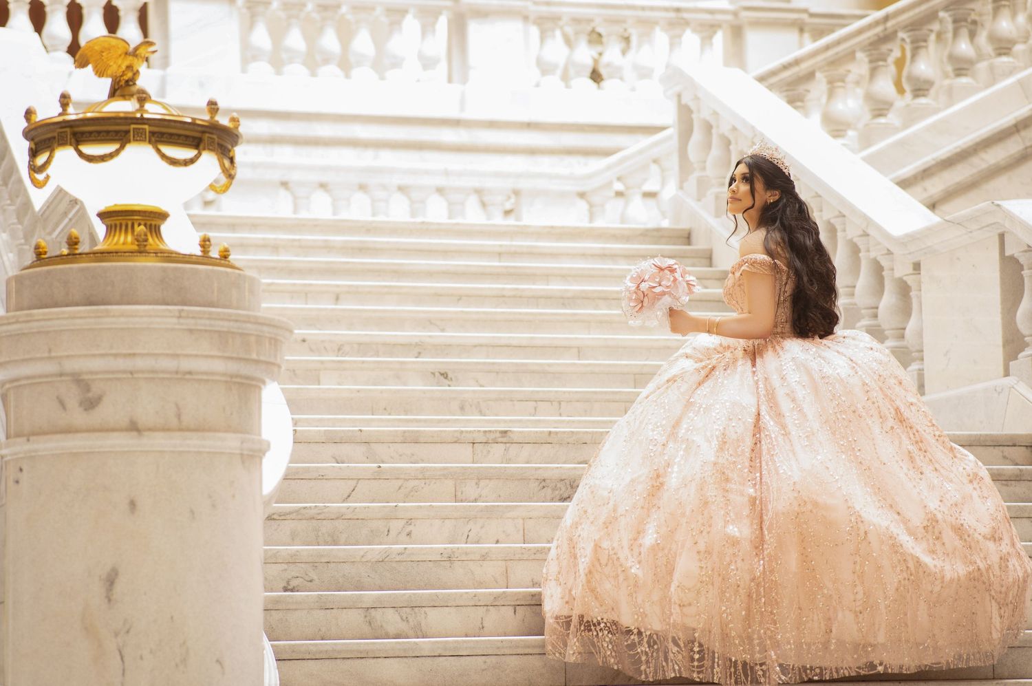 Elegant bride in a pink ball gown posing on marble stairs at a grand venue.