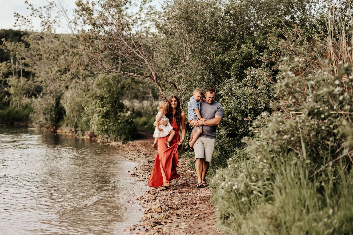 Family walking along a scenic river bank surrounded by lush greenery on a sunny day.