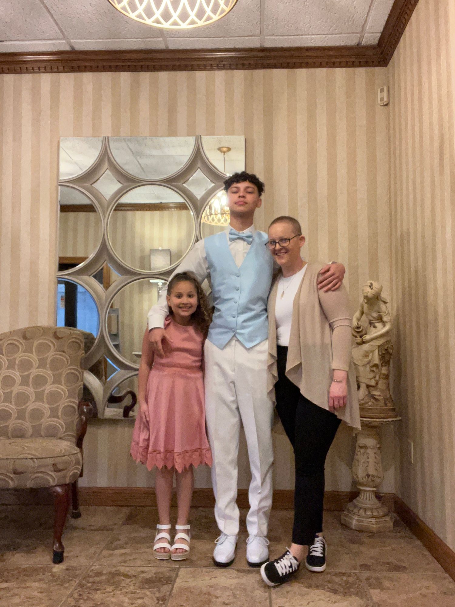 Three people pose together in formal attire against a decorative wall with mirrored panels and elegant decor.