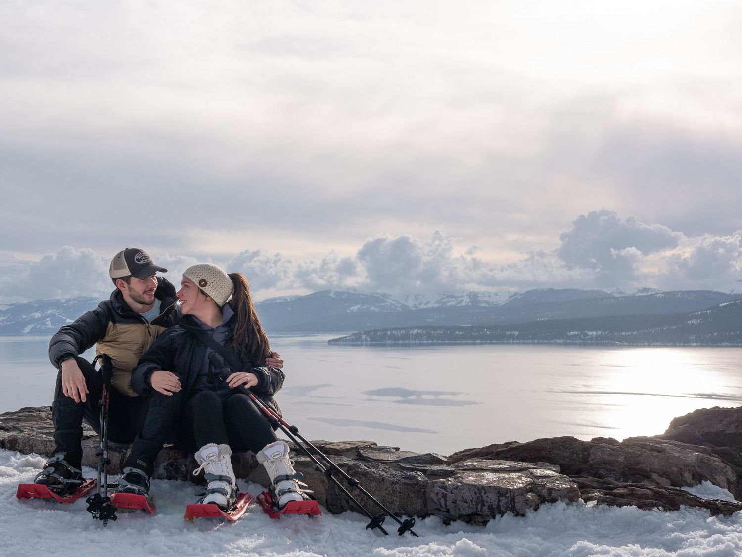 A couple sits together on snowy rocks overlooking a scenic mountain lake vista.