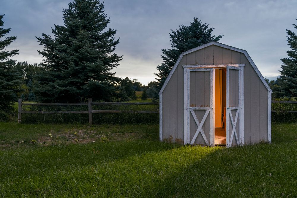 Illuminated barn-style storage shed at dusk with pine trees in background.