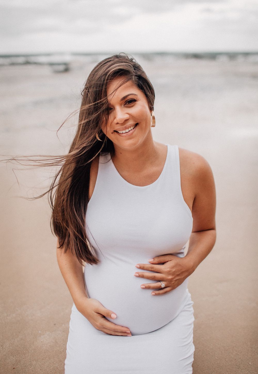 Maternity photos captured at the beach with a woman in white dress touching her baby bump at sunset.