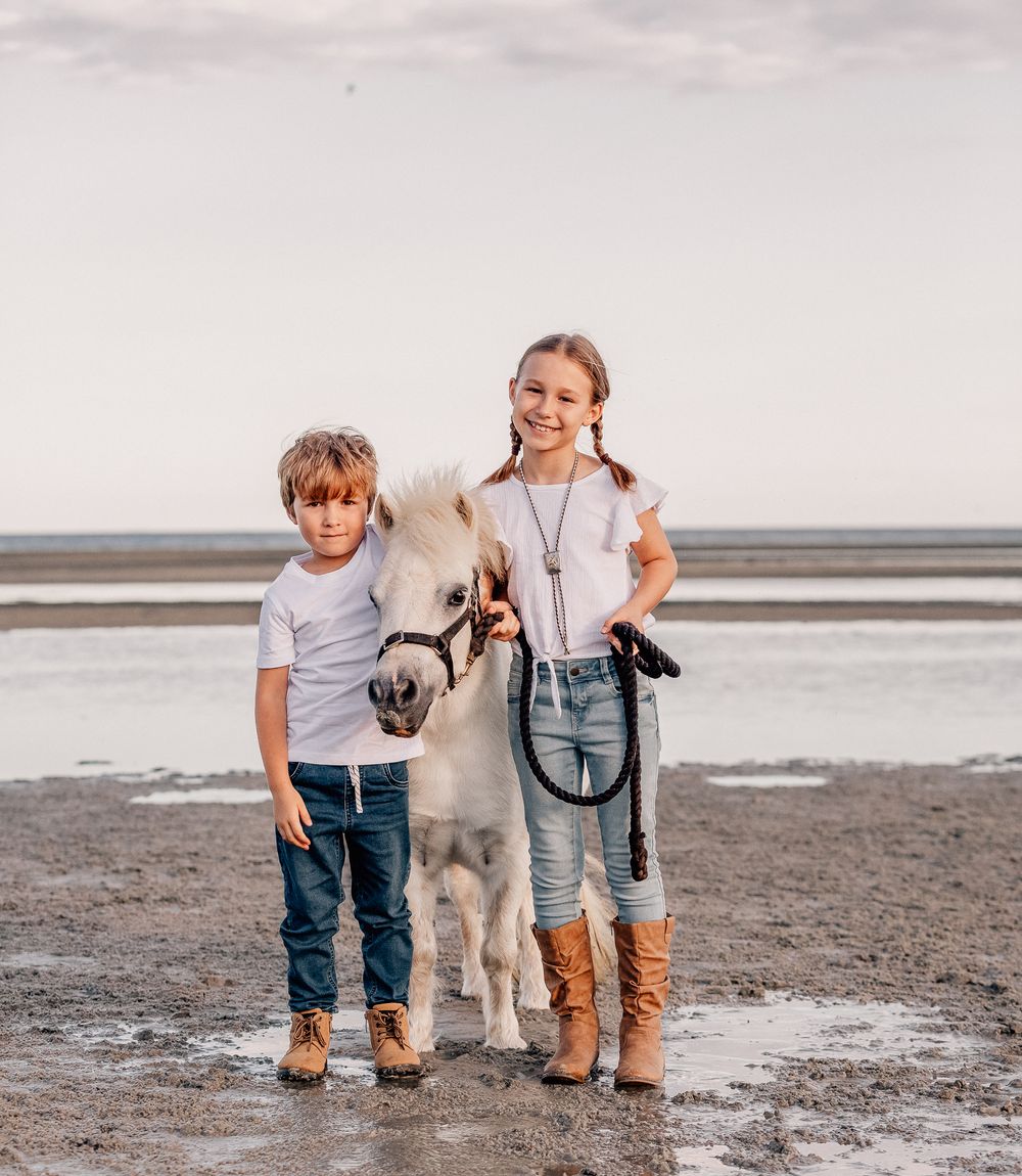 Two children standing with a white pony on a beach during sunset.
