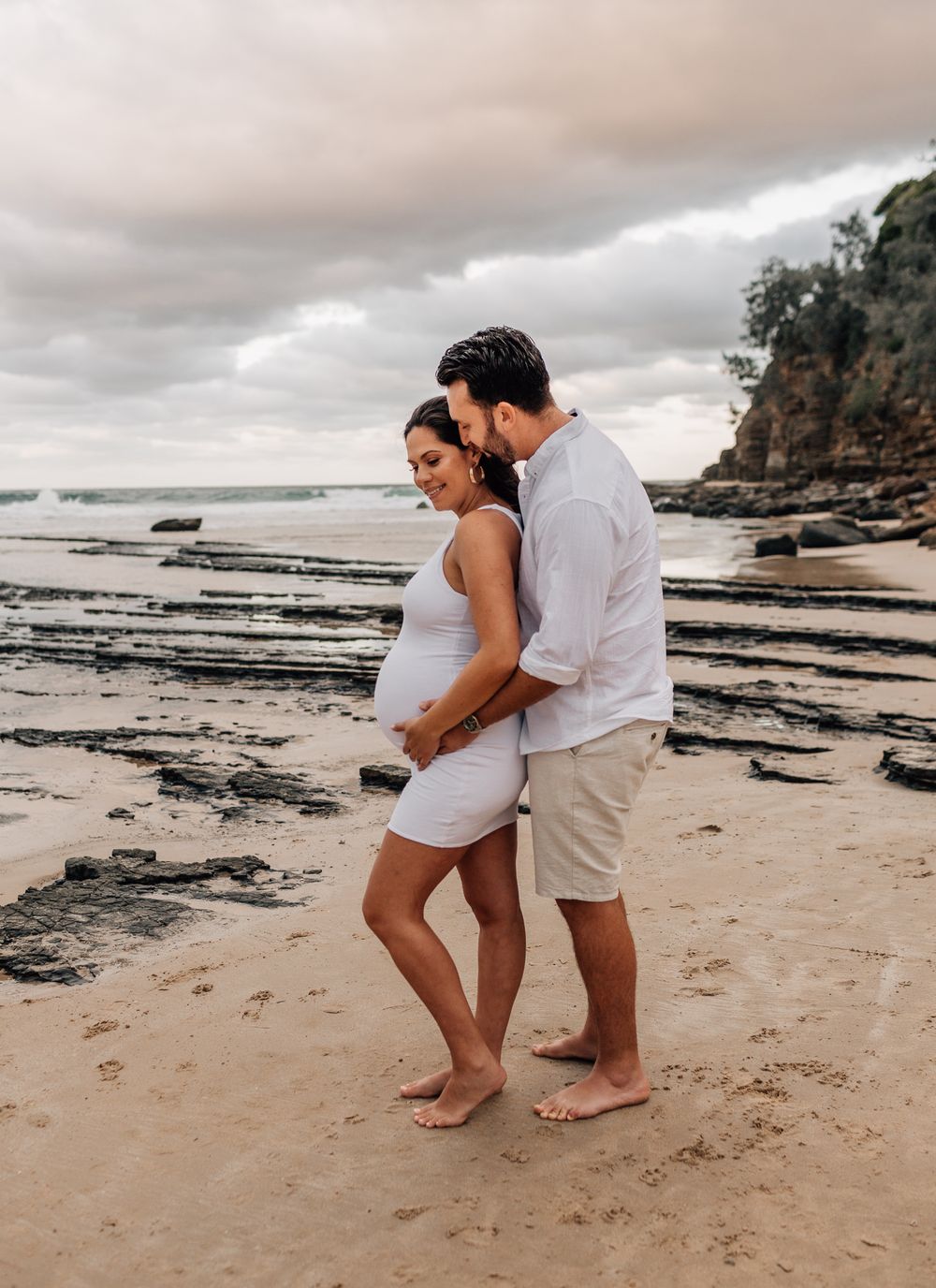 Romantic beach maternity photos with couple walking on sand at sunset.