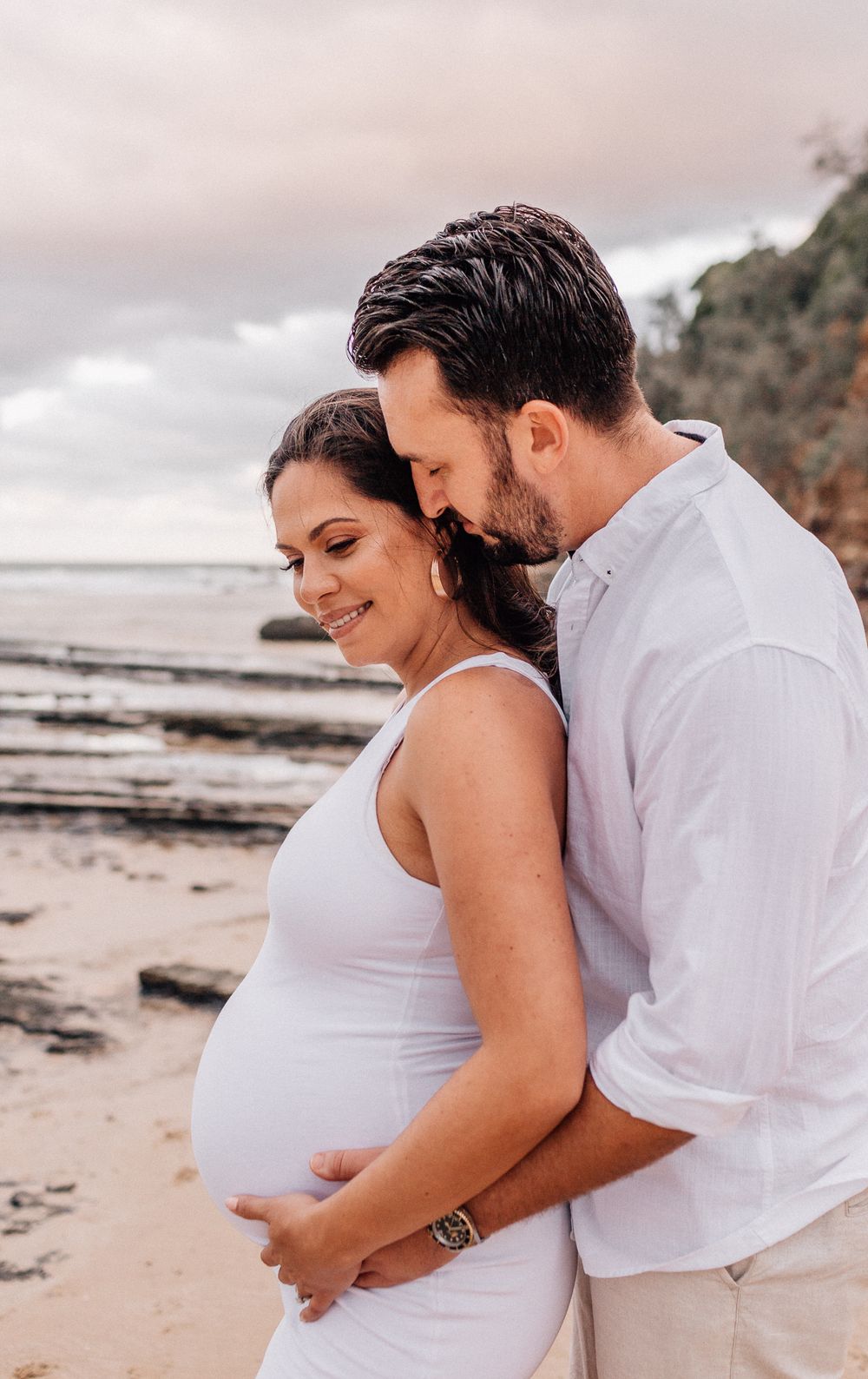 Maternity photoshoot on beach showing couple in white embracing.
