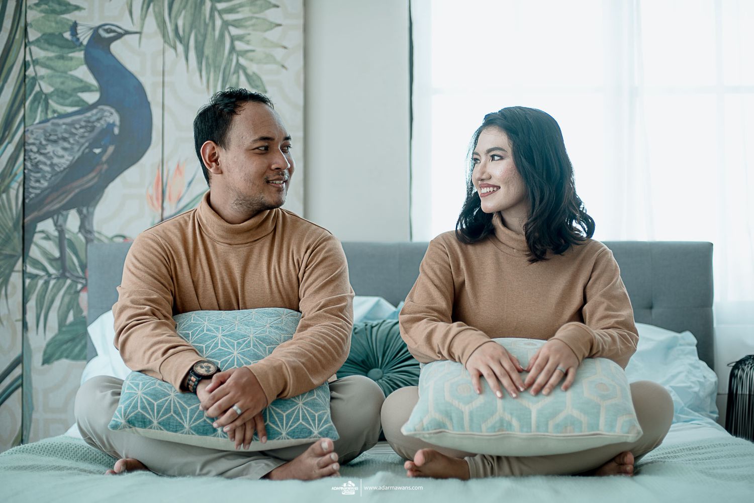 A couple in matching beige sweaters sits on a couch with tropical wall art.