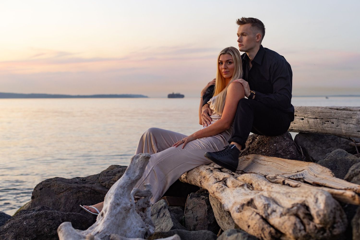 Couple sits romantically on coastal rocks at sunset overlooking a calm ocean view.