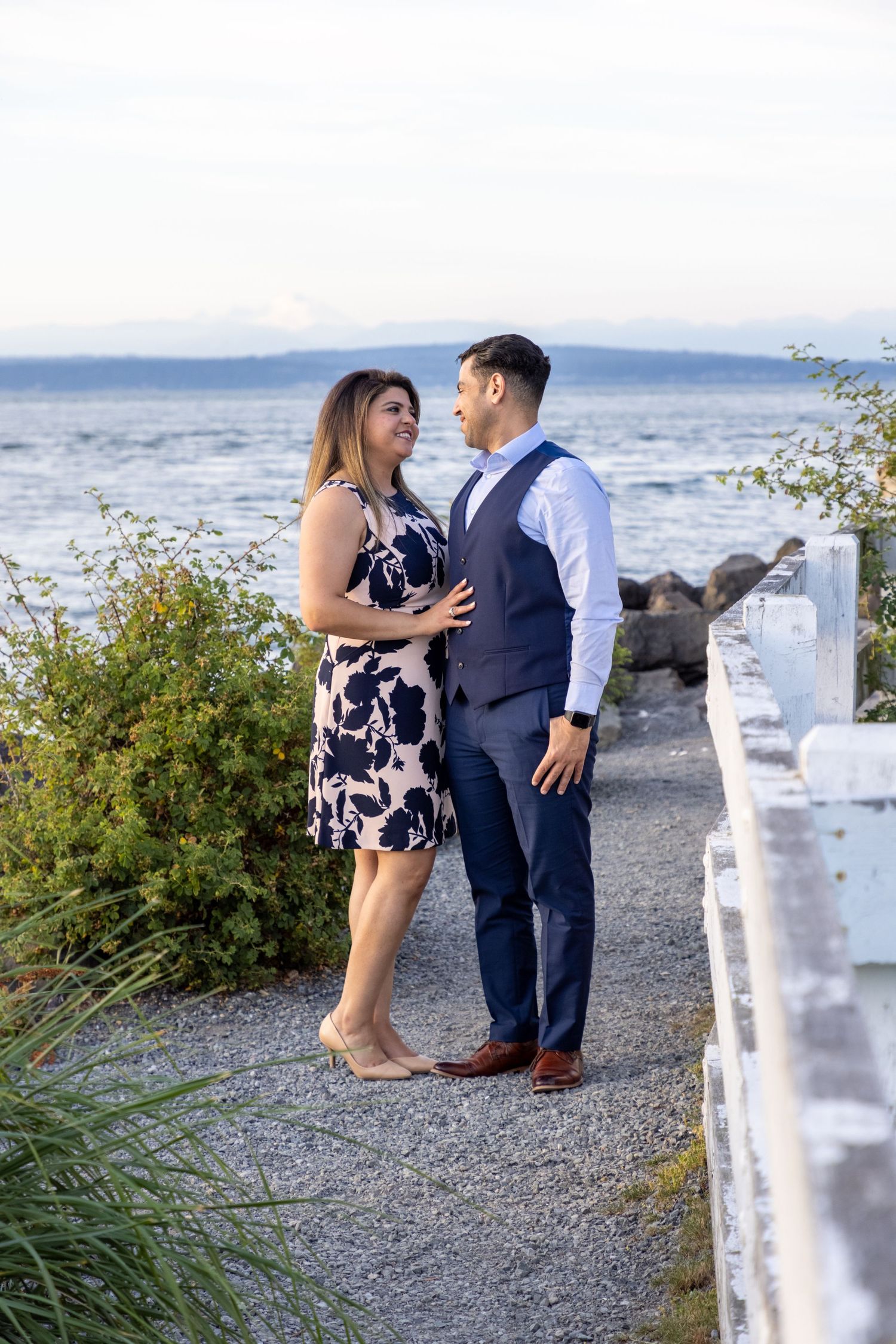 Couple poses along a seaside path with white fence and ocean view in the background.