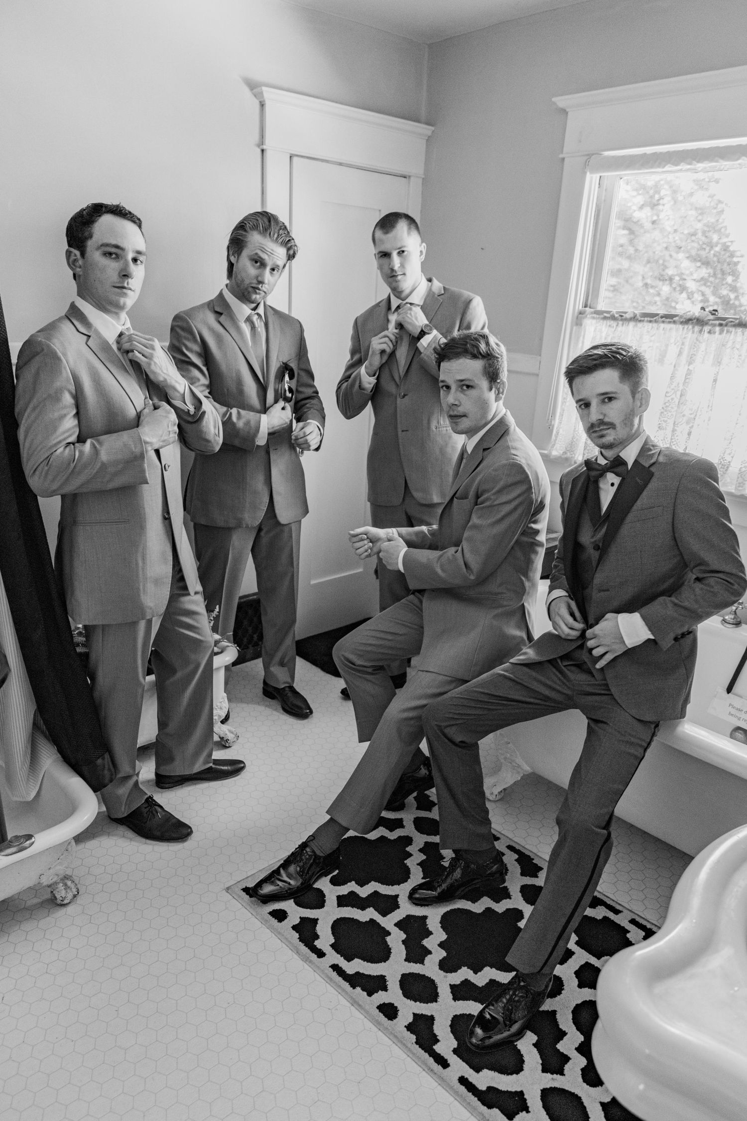Group of groomsmen in formal suits pose together in bright indoor room.