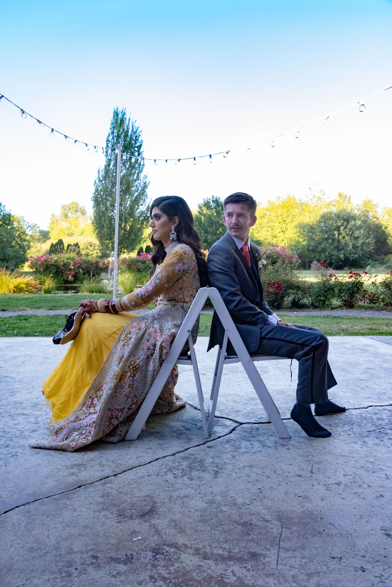 Couple sits back-to-back on white chairs at outdoor reception with string lights.