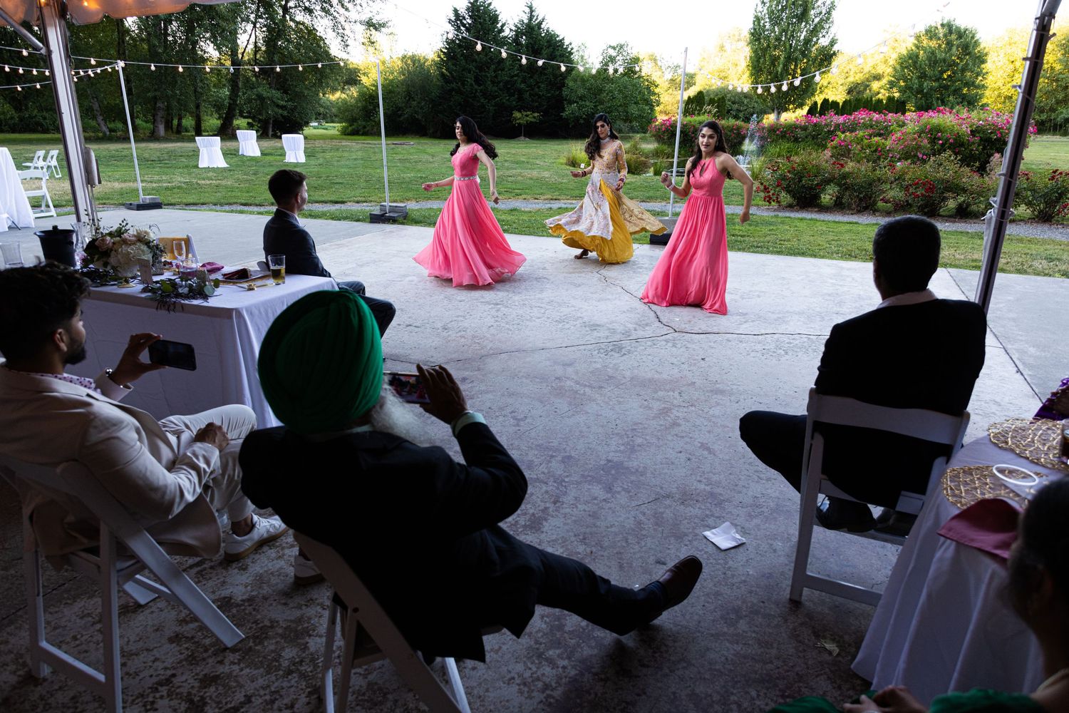 Wedding guests watch performers in pink dresses dancing during an outdoor reception.