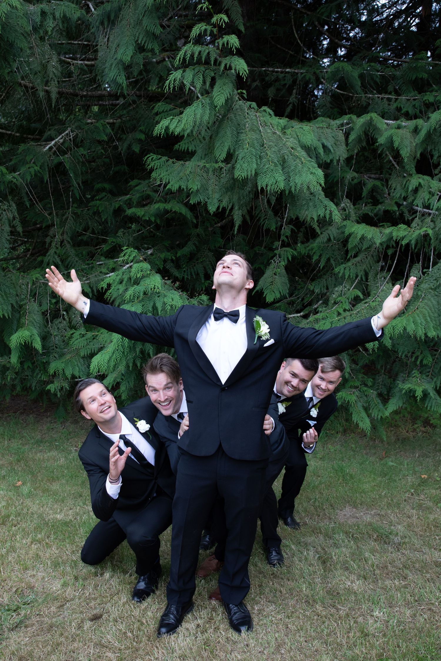 Groomsmen strike a humorous pose behind the groom with arms spread wide against evergreen trees.