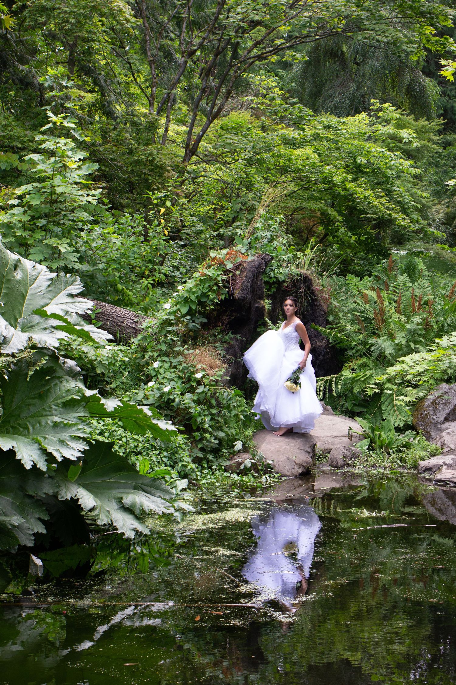 A bride in a white dress poses gracefully by a pond in a lush garden setting.