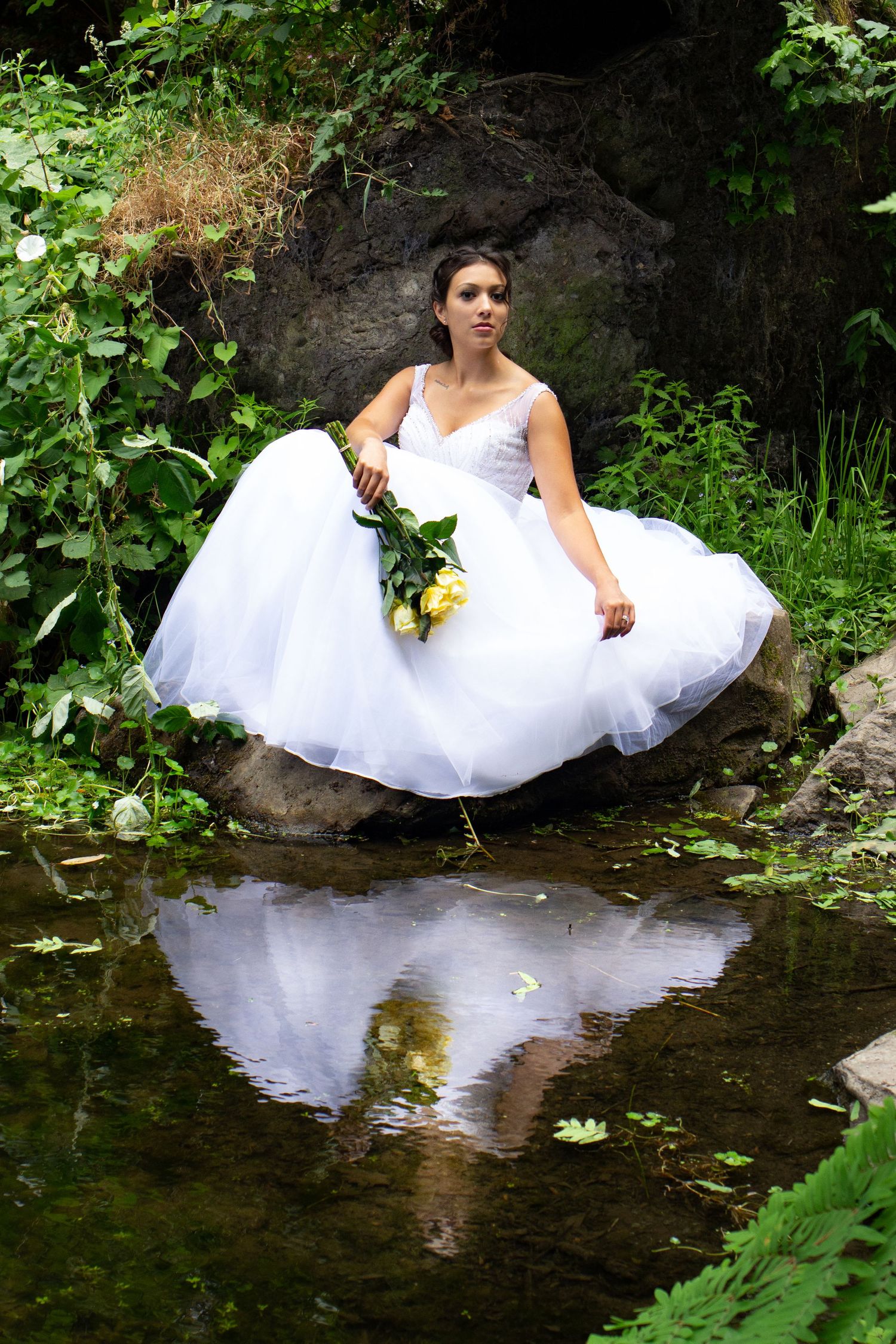 A bride in a white tulle gown sits on rocks by a reflective pond surrounded by greenery.
