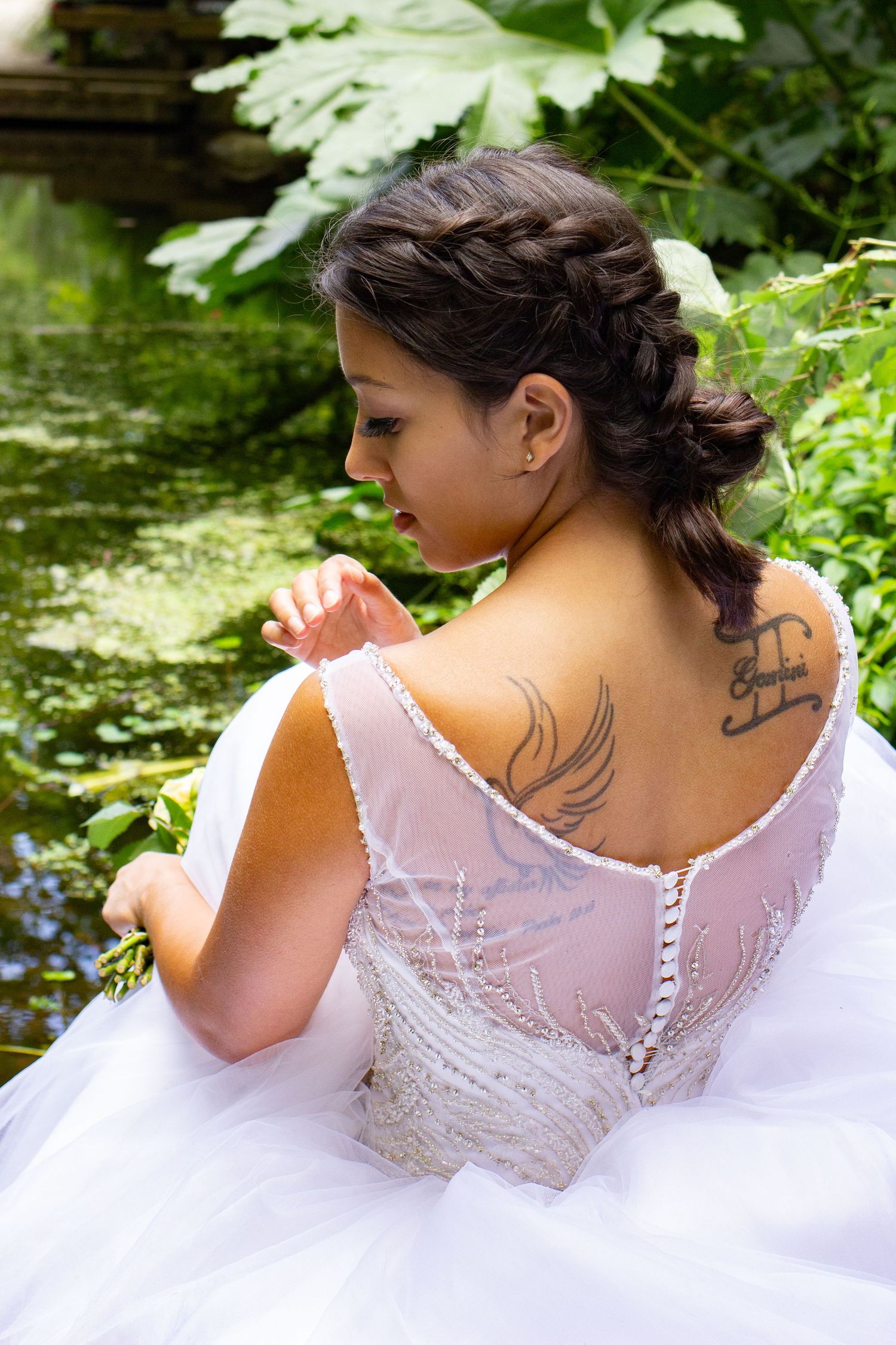 A white wedding dress with a detailed lace back and braided hairstyle captured near a pond.
