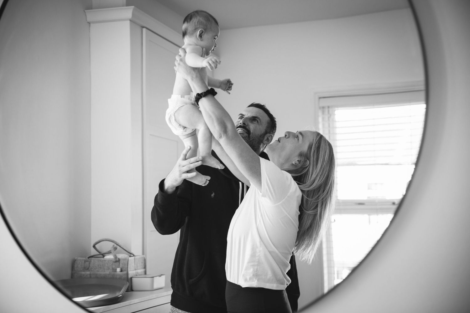 Black and white mirror reflection captures playful moment lifting baby in the air.