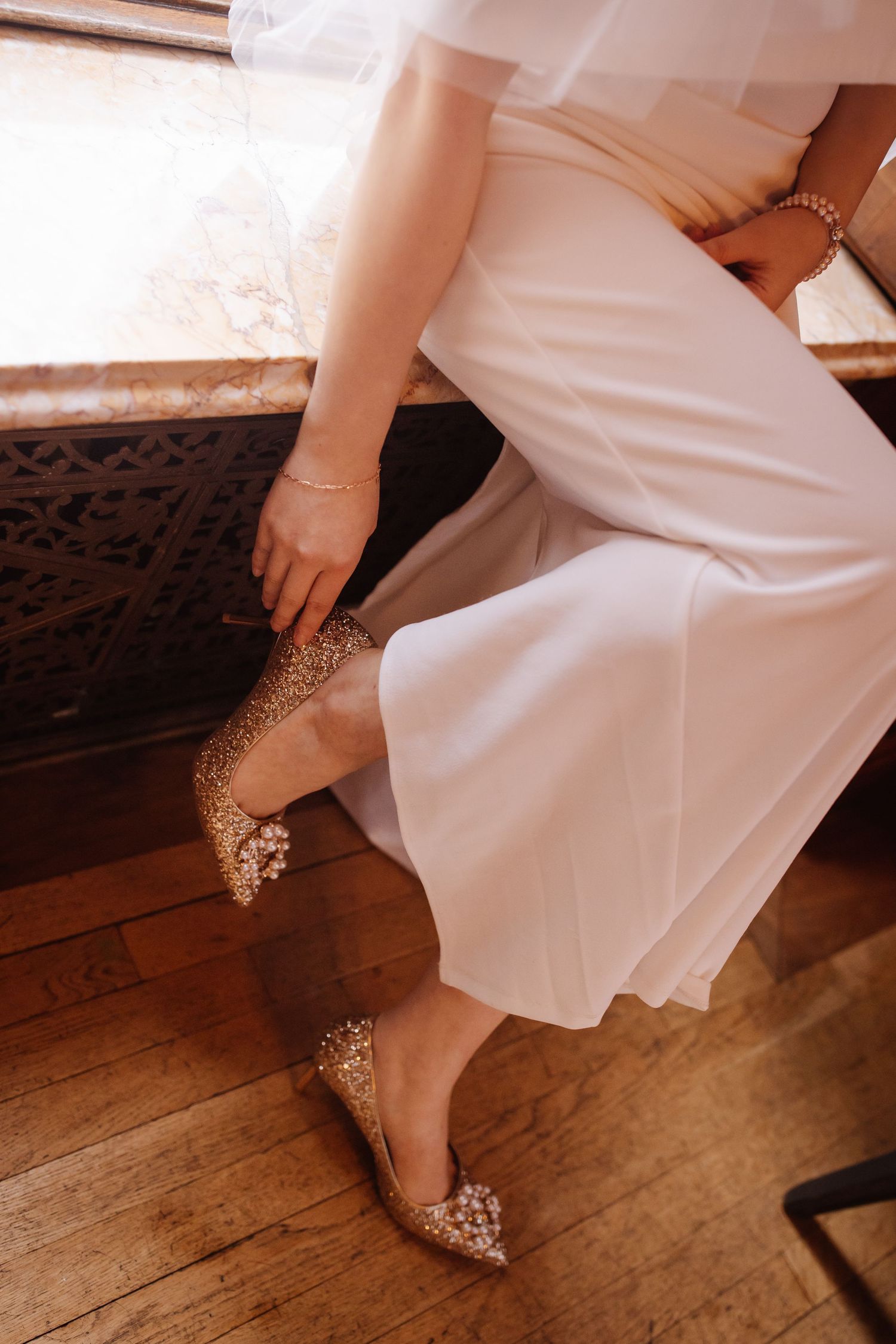 Sparkly bridal shoes and white dress hem detail on hardwood floor.