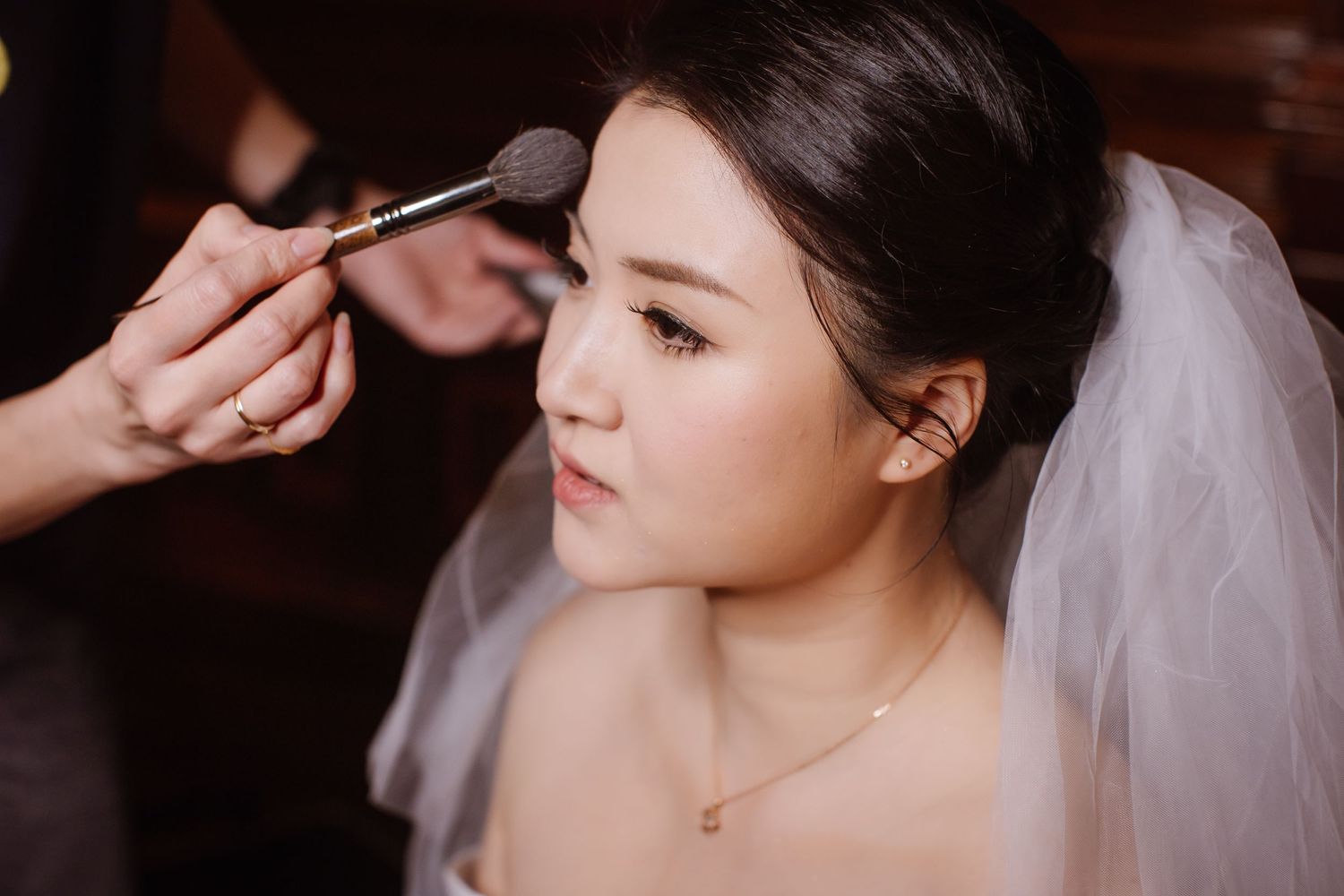 Makeup application in progress with white bridal veil and delicate necklace.