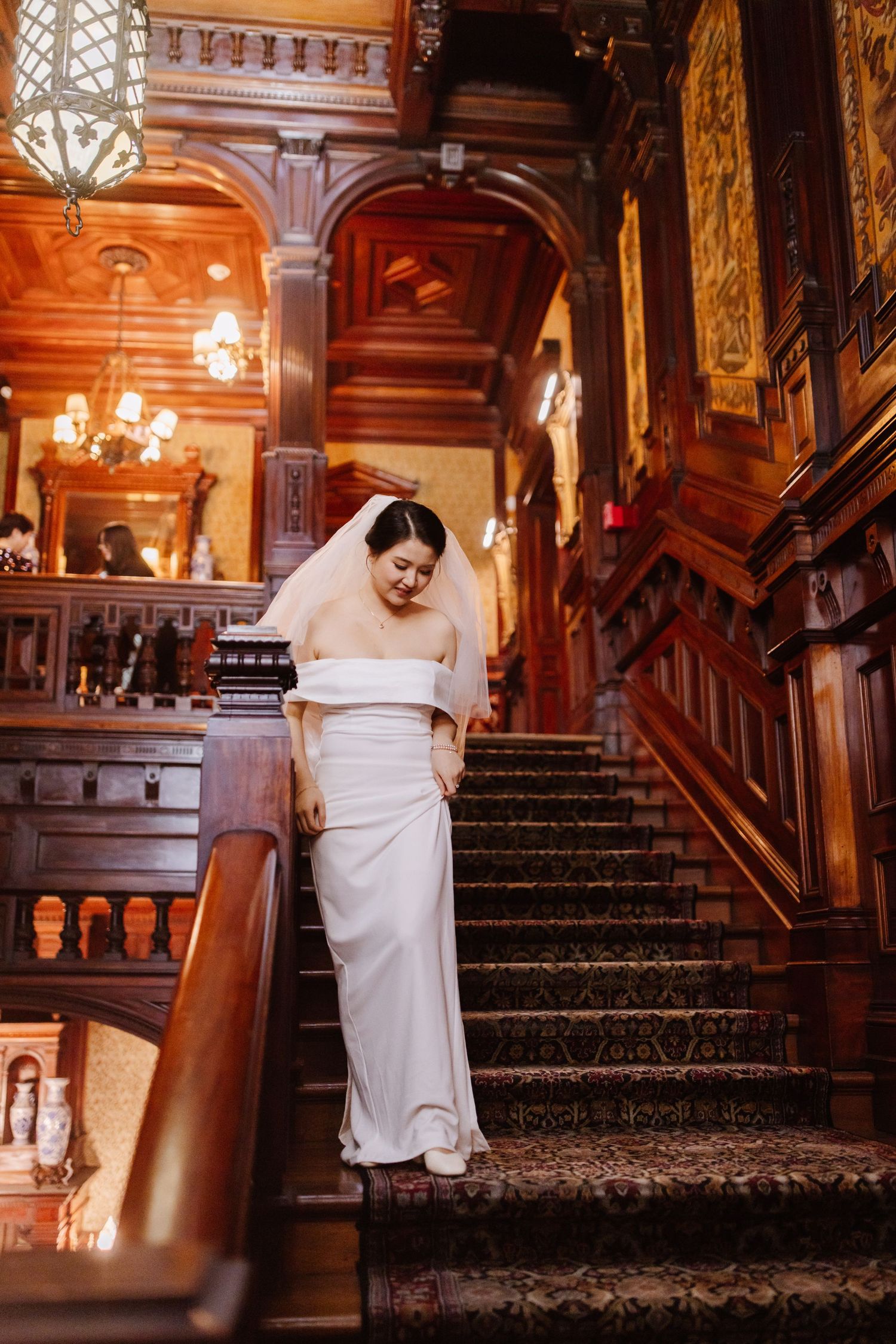 Elegant bride descending ornate wooden staircase in vintage mansion.