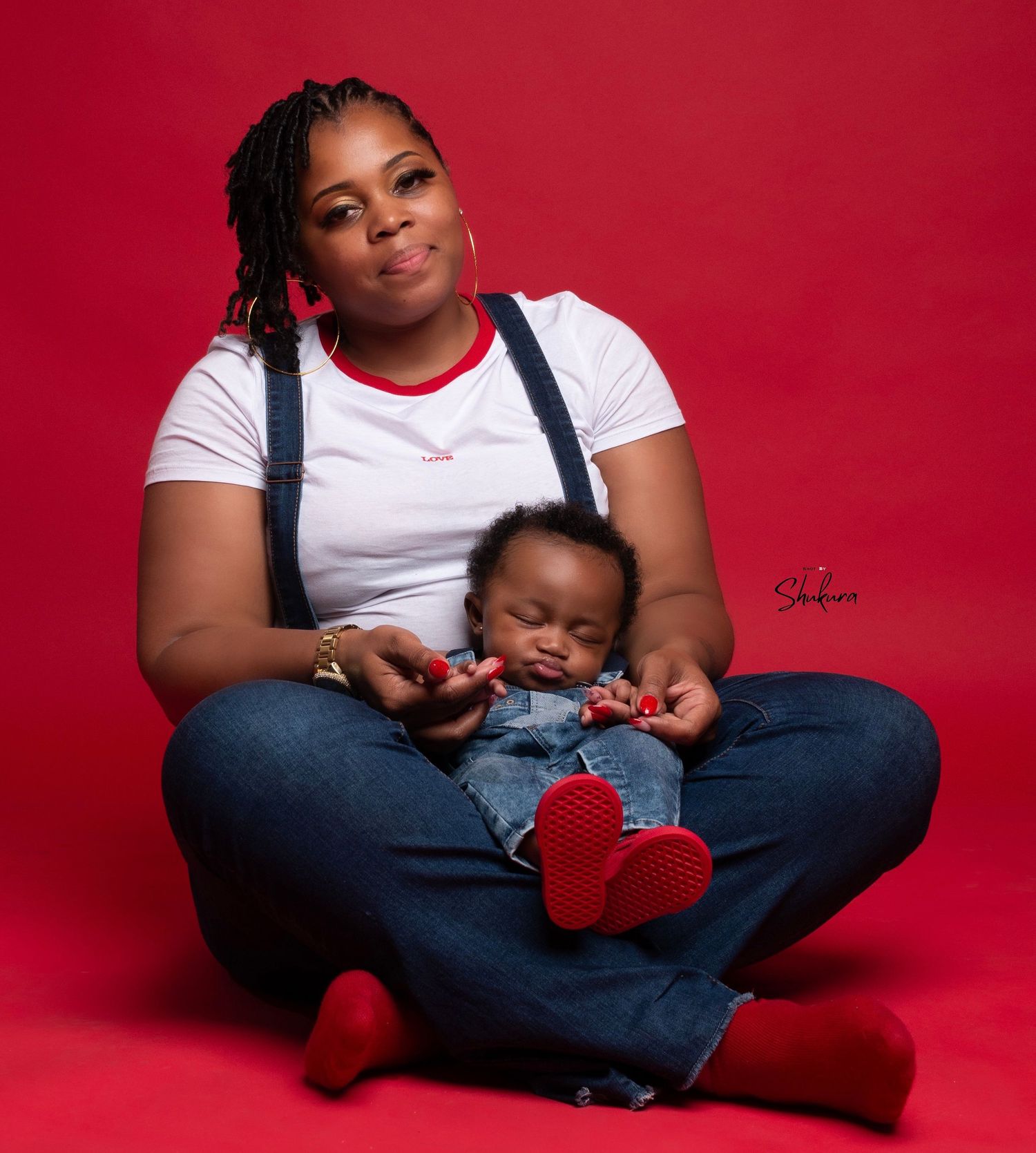 Parent in overalls cradling sleeping baby against red backdrop.