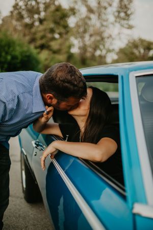 A couple shares a romantic moment leaning into a vintage blue car during a photoshoot.
