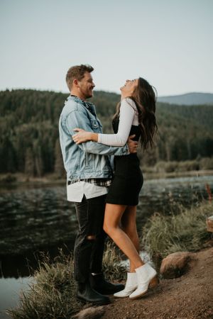 Couple in casual wear sharing playful moment on hillside trail overlooking lake.
