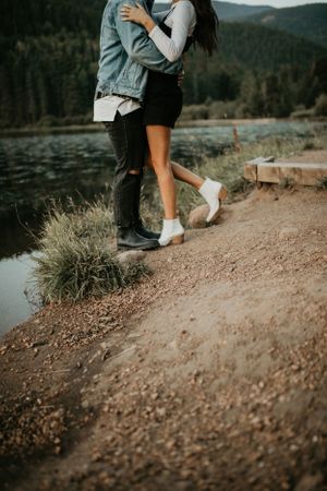 A couple embraces on a rocky lakeside trail with mountains in the background during golden hour.