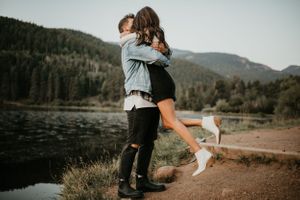 Candid engagement photography session on mountain trail with lake and forest backdrop.