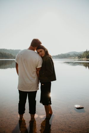 Silhouetted couple stands together at edge of serene lake during moody photoshoot.
