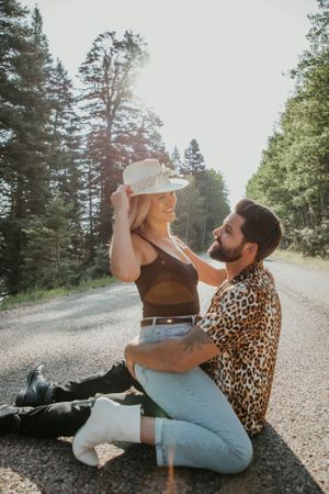Two people sitting playfully on a dirt road surrounded by evergreen trees.