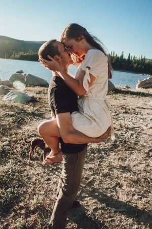 Romantic couple embraces on a rocky lakeshore with mountains in the background.