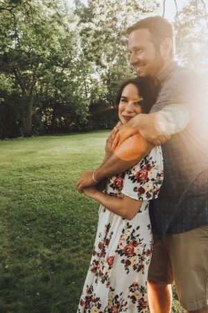 Couple embracing in a sunlit garden with a floral dress against green scenery.