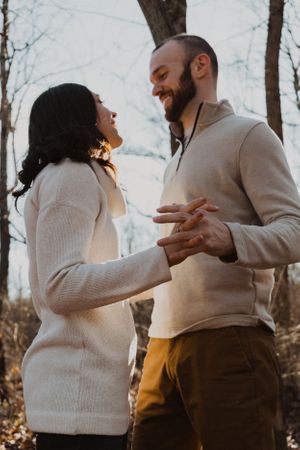 A couple in winter clothing embraces and laughs together in a wooded setting.
