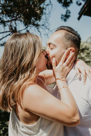 A romantic couple kisses during a bright sunny outdoor photoshoot with trees in background.