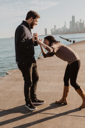 Playful couple dancing and laughing together on a lakefront with city skyline in background.