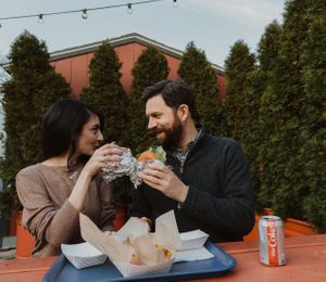 Couple enjoying takeout food and drinks at outdoor table with string lights.